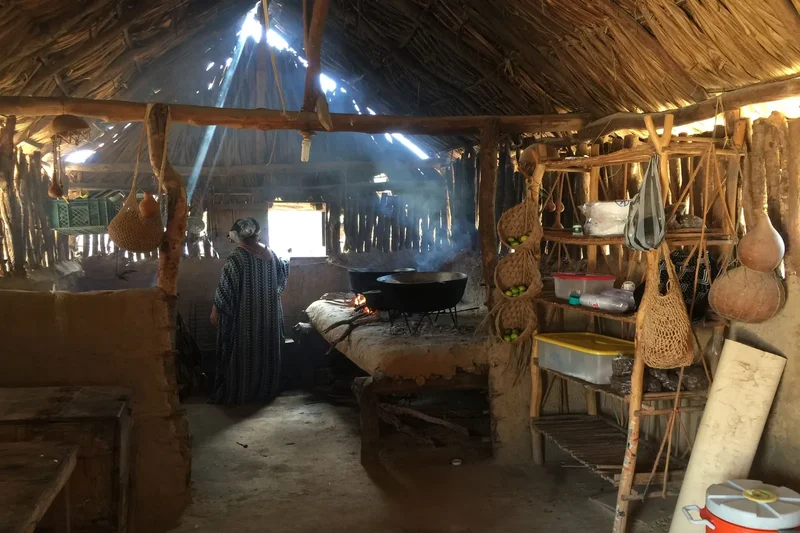 Interior de cocina en ranchería wayuu con fogón de leña y mujer mayor preparando alimentos